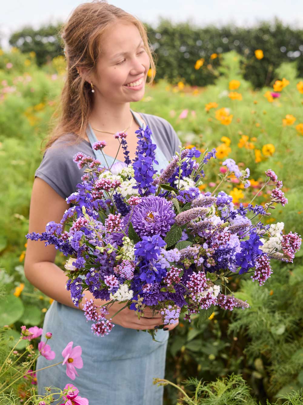 A smiling woman standing in a flower field holds a large bouquet of purple, lavender, and white wildflowers.