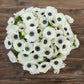 A boquet of white anemone flowers with dark centers and green foliage on a wooden table.