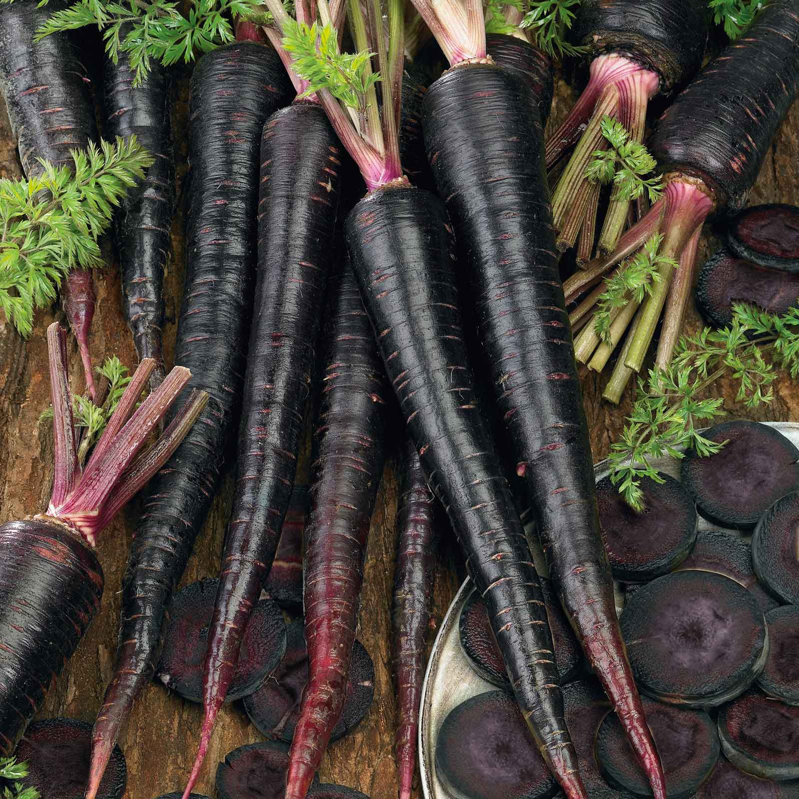 A group of black nebula carrots showing the nearly black skin with slices of the carrot surrounding it on a wooden surface.
