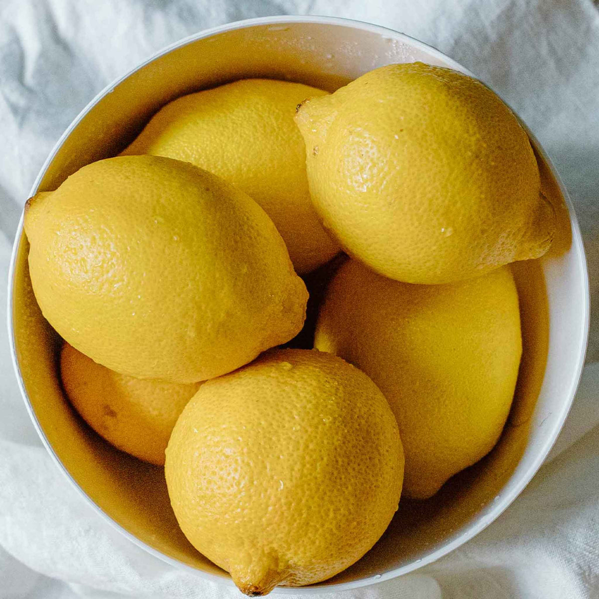 Several bright yellow lemons from the meyer lemon bush arranged in a white bowl on a light fabric surface.