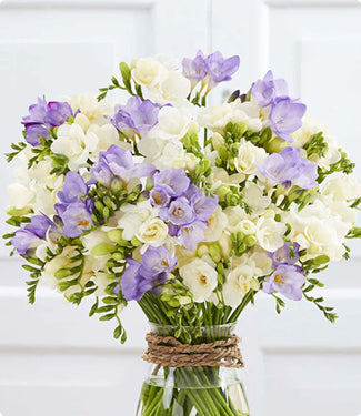 A vibrant flower bouquet featuring white and light purple flowers, arranged in a clear glass jar with a rustic twine wrap, set against a clean white paneled background.