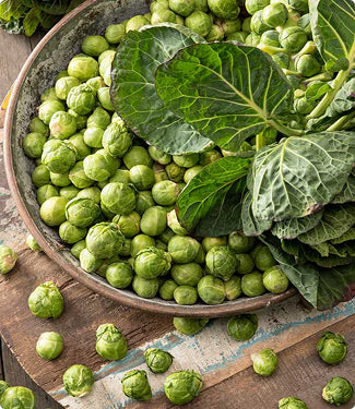 A rustic bowl filled with green Brussels sprouts and large leafy greens on a wooden surface.